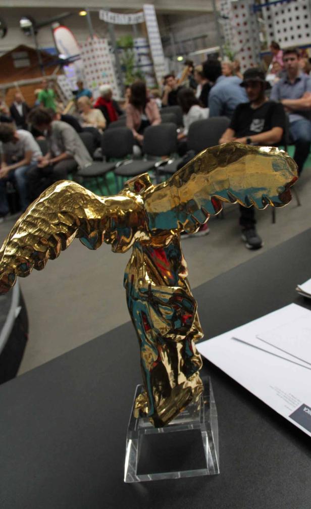 Eine goldene Statue der Siegesgöttin Nike steht auf einem Tisch vor Publikum.