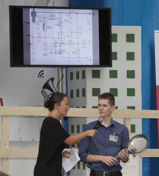 Eine Frau interviewt einen jungen Mann mit einem Mikrofon vor einem Schaltplan.