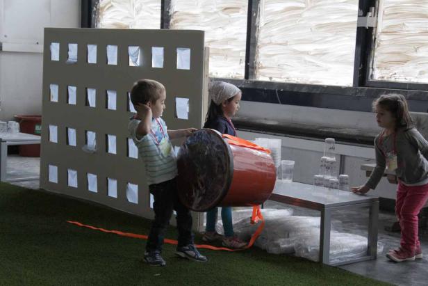 Drei Kinder spielen mit einem großen Fass und Plastikbechern in einem Innenraum.