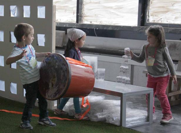 Drei Kinder spielen mit einem Fass und Plastikbechern.
