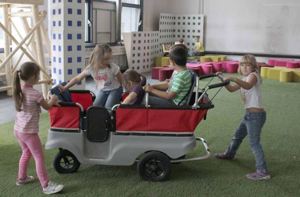 Mehrere Kinder spielen mit einem Bollerwagen in einem Innenraum.