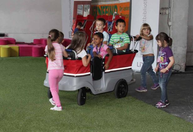 Eine Gruppe Kinder spielt mit einem kleinen, roten Wagen auf einer künstlichen Grasfläche.