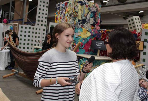 Eine junge Frau interviewt eine andere Person mit einem Mikrofon vor einem farbenfrohen Hintergrund.