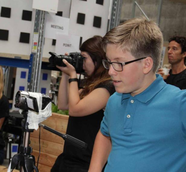 Ein Jugendlicher steht vor einer Frau mit einer Kamera und einer Videokamera auf einem Stativ.