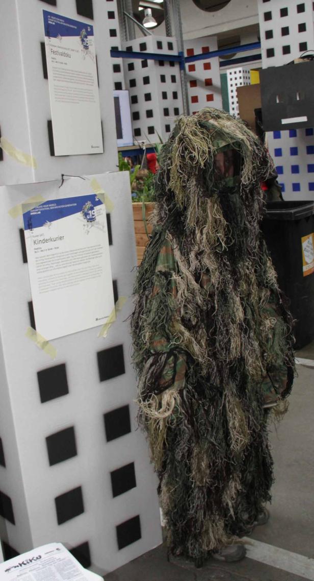 Eine Person in einem Ghillie-Anzug steht neben einem Stand mit Flyern.
