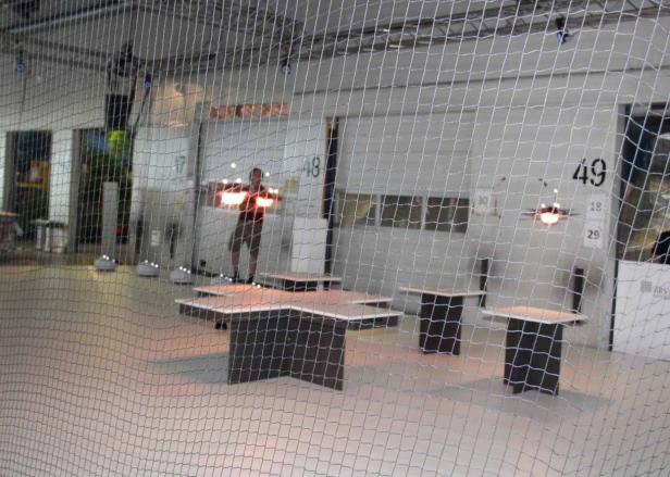 Ein Mann steuert eine Drohne in einer Halle mit Tischen und einem Netz im Vordergrund.