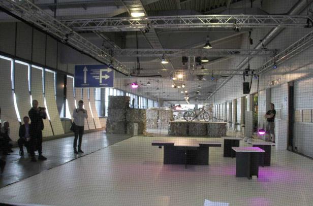 Eine Ausstellungshalle mit Installationen aus recyceltem Material und einem Fahrrad auf einem Podest.