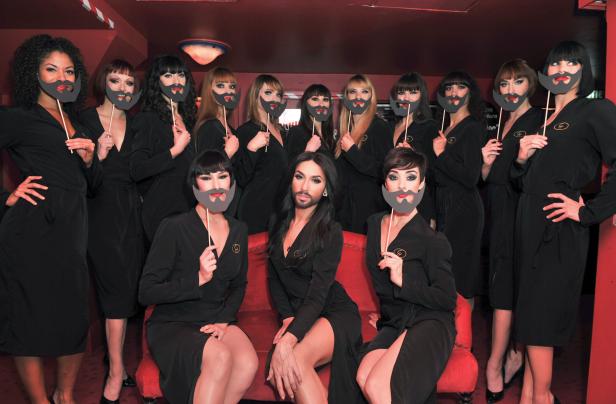 Dragqueens posieren mit aufgeklebten Bärten in einem rot beleuchteten Raum.
