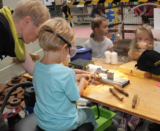 Ein Kind arbeitet mit Schutzbrille an einer Drehbank unter Aufsicht eines Erwachsenen.