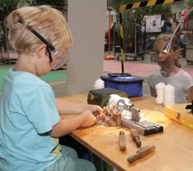 Ein Junge mit Schutzbrille arbeitet an einer kleinen Drehbank, während ein Mädchen zusieht.
