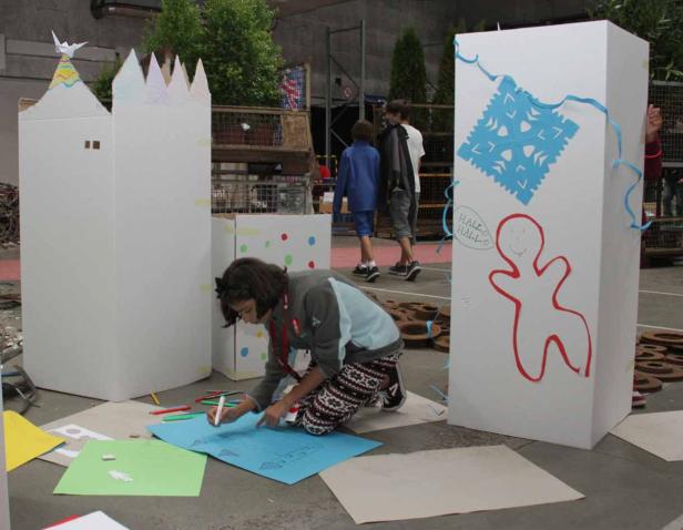 Ein Mädchen zeichnet auf blauem Papier vor bemalten Kartonbauten.