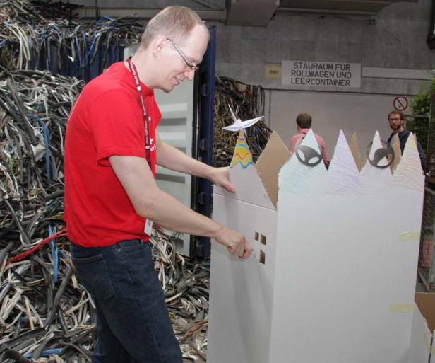 Ein Mann baut an einem Modellhaus aus Pappe vor einem Container mit Kabelresten.