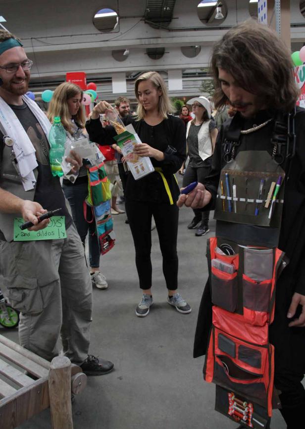 Mehrere Personen stehen in einem Raum, einige mit ungewöhnlichen Accessoires wie Werkzeugwesten und Spielzeugwaffen.