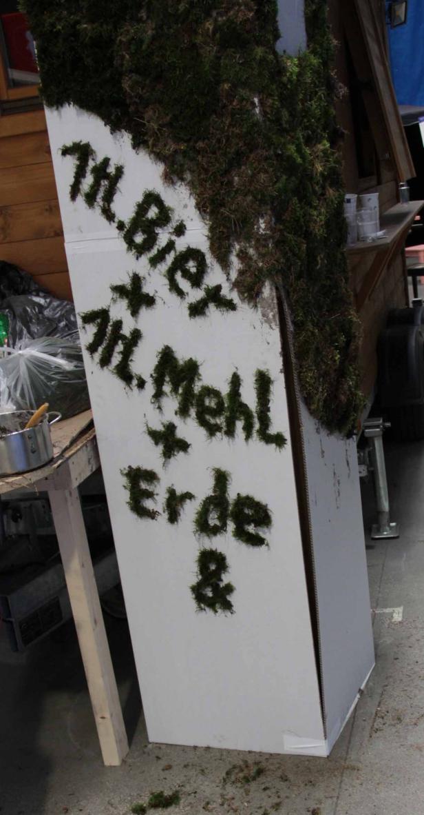 Auf einer weißen Fläche steht mit Moos geschrieben: „H.Bier + H.Mehl + Erde &“.