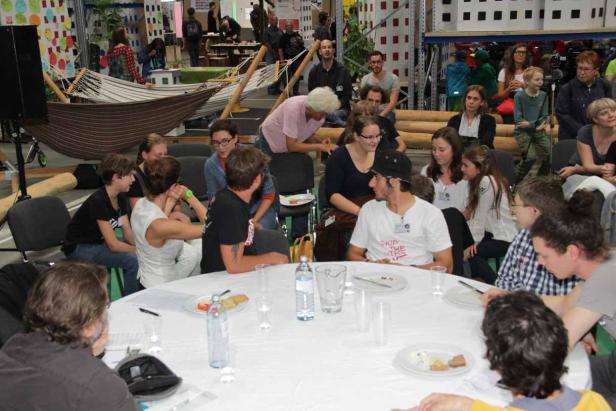 Menschen sitzen an einem runden Tisch und schauen einer Präsentation in einem Lagerhaus zu.