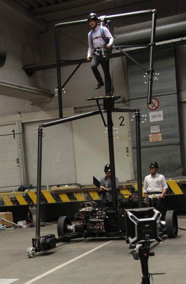 Ein Mann steht auf einer Plattform, die Teil einer großen, fahrbaren Apparatur ist. Zwei weitere Männer beobachten ihn.