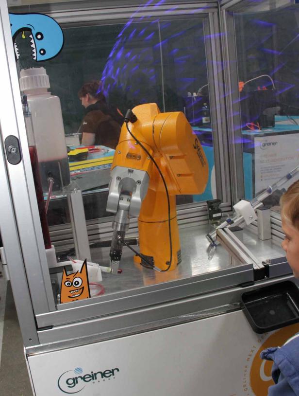 Ein gelber Roboterarm steht in einem Glaskasten mit bunten Aufklebern.