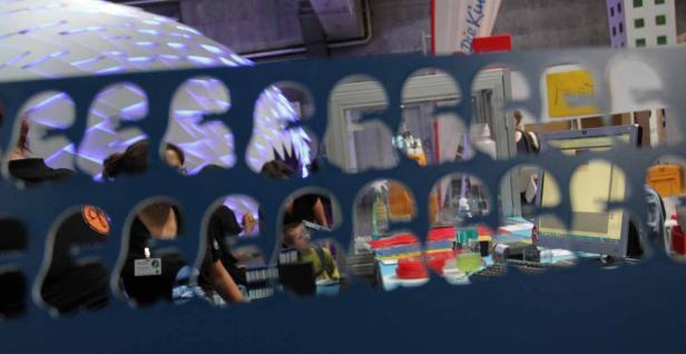 Ein Blick durch eine Lochblende auf einen Messestand mit Menschen und Computern.