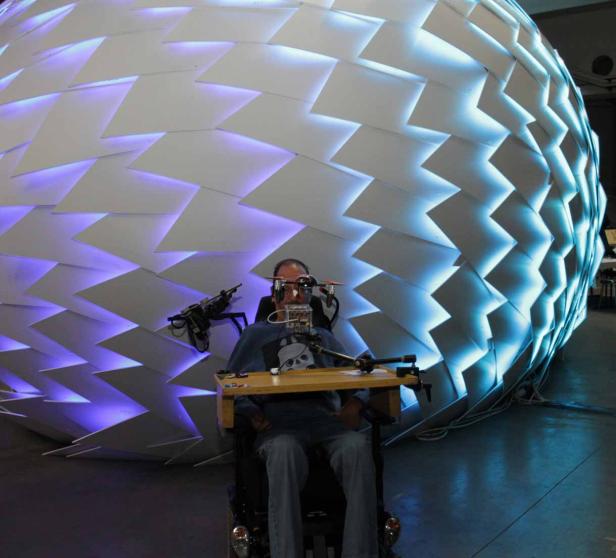 Ein Mann im Rollstuhl mit einer technischen Vorrichtung vor einem großen, beleuchteten Objekt.