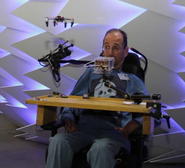 Ein Mann im Rollstuhl steuert eine Drohne mit einer Vorrichtung am Mund.