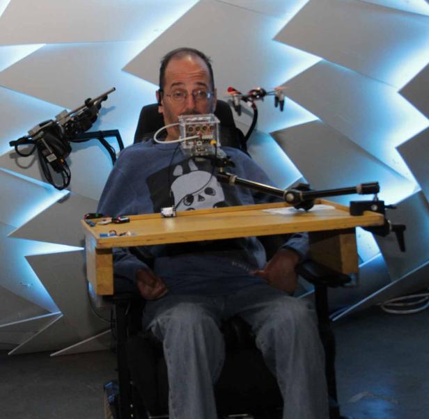 Ein Mann im Rollstuhl mit einer speziellen Vorrichtung vor dem Mund sitzt vor einer Wand mit geometrischen Formen.