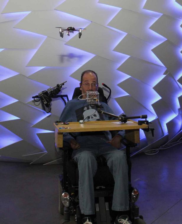 Ein Mann im Rollstuhl steuert eine Drohne mit einer Vorrichtung vor seinem Mund.