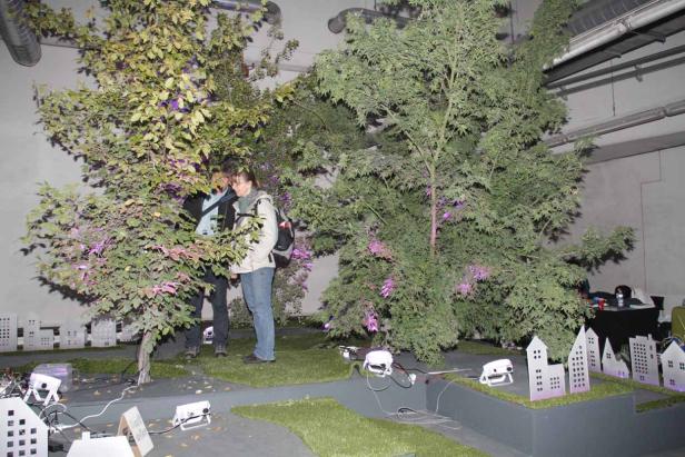 Zwei Personen betrachten eine Installation mit Bäumen und Modellhäusern in einem Innenraum.