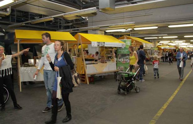 Menschen besuchen einen Markt mit gelben Ständen in einer Halle.