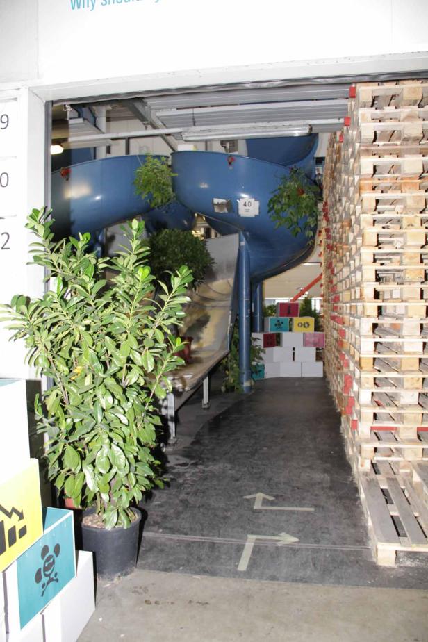 Eine blaue Rutsche schlängelt sich durch einen Lagerraum, dekoriert mit Pflanzen.