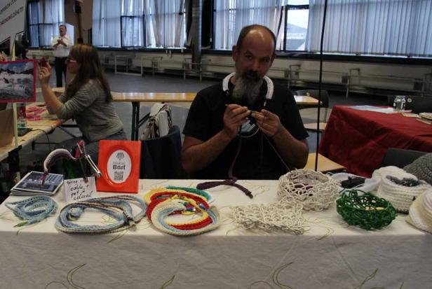Ein Mann mit Bart häkelt an einem Stand bunte Kabelummantelungen.