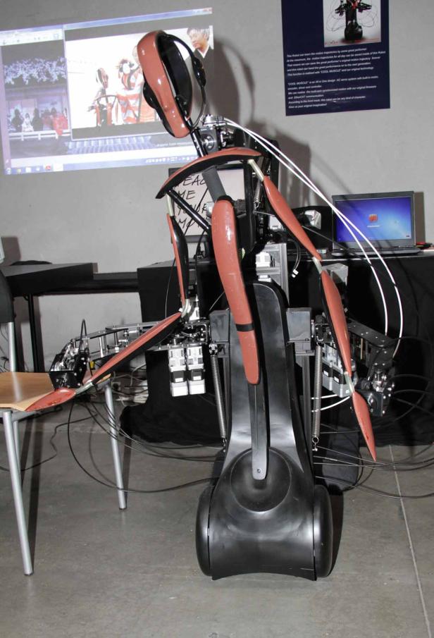 Ein humanoider Roboter mit braunen Akzenten steht in einem Raum mit einem projizierten Bild an der Wand.