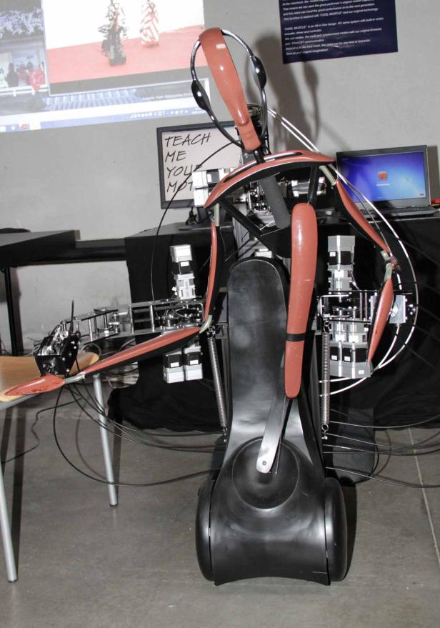 Ein komplexer Roboter mit Kopfhörern steht in einem Raum mit einem Laptop und einem Whiteboard.