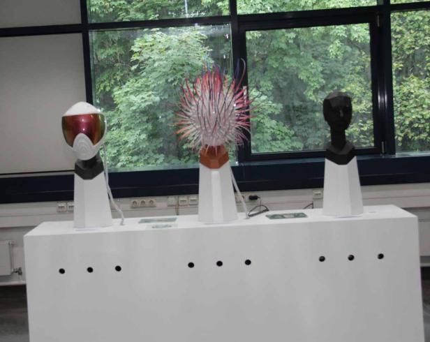 Drei verschiedene Skulpturen stehen auf einem weißen Sockel vor einem Fenster.