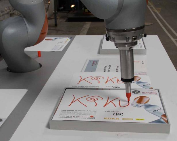 Ein Roboterarm schreibt mit einem roten Stift das Wort „KUKA“ auf eine Verpackung.