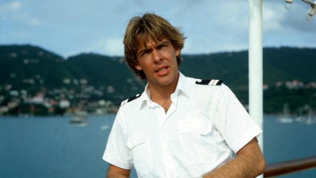 Ein Mann in Kapitänsuniform steht an Bord eines Schiffes vor einer tropischen Landschaft.