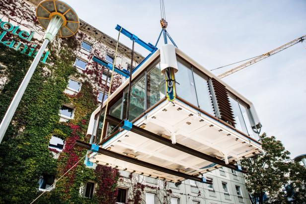 Ein vorgefertigter Raum wird mit einem Kran vor einem mit Efeu bewachsenen Hotel platziert.