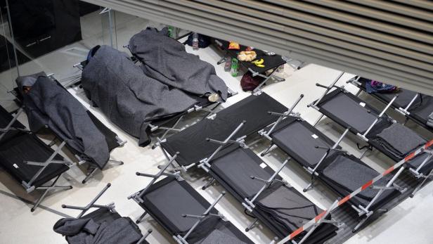 Mehrere Feldbetten stehen in einem Raum, einige sind mit schlafenden Personen unter grauen Decken belegt.