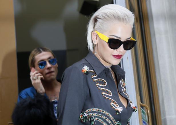 Rita Ora mit blonden Haaren und Sonnenbrille vor einem Gebäude.