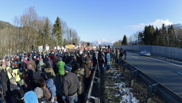 Eine Menschenmenge demonstriert neben einer Autobahn mit schneebedeckten Bergen im Hintergrund.