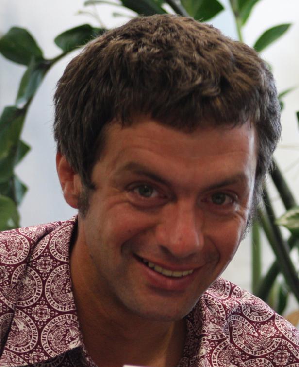 Ein lächelnder Mann mit gemustertem Hemd vor einer Zimmerpflanze.