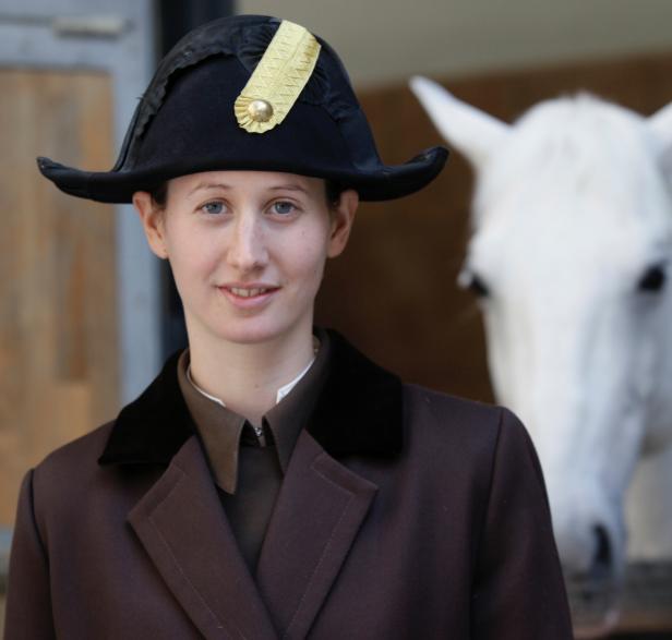 Eine Frau mit Dreispitz und braunem Reitrock steht vor einem weißen Pferd.