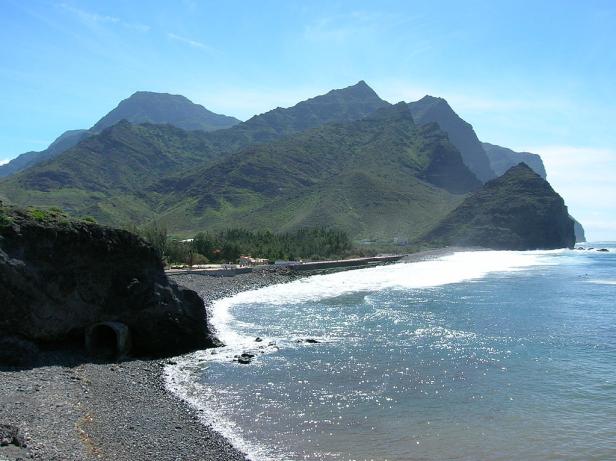 Blick auf einen Strand mit dunklem Sand und dahinterliegenden Bergen.