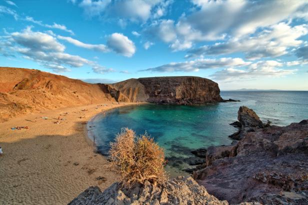 Blick auf einen Strand mit türkisfarbenem Wasser und roten Felsen unter blauem Himmel mit Wolken.