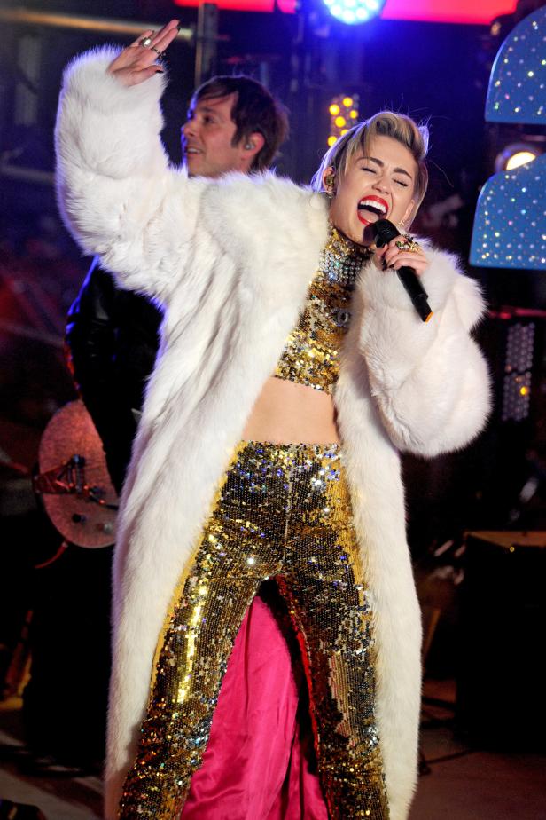 Miley Cyrus tritt in einem glitzernden Outfit und einem weißen Pelzmantel auf.