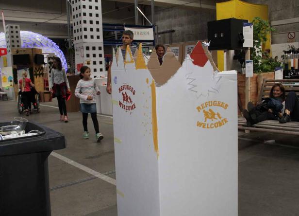 Kinder und Erwachsene in einer Halle mit einer Installation „Refugees Welcome“.