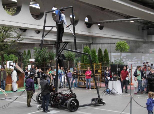 Eine Person steht auf einer großen, selbstgebauten Maschine mit Rädern, umgeben von Zuschauern.