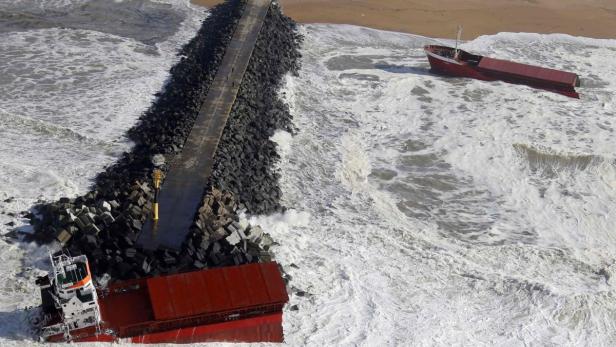 Ein gestrandetes Schiff liegt neben einer Mole im aufgewühlten Meer.