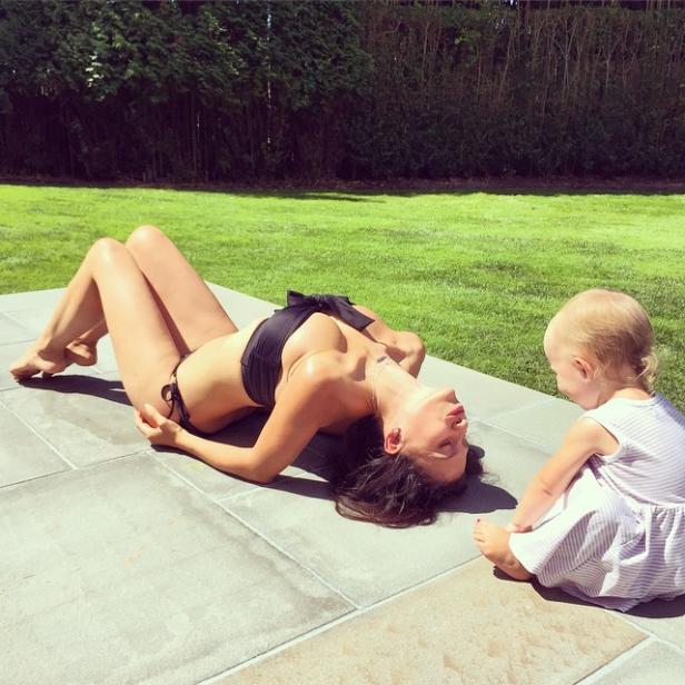 Eine Frau im Bikini liegt auf einer Terrasse, während ein kleines Kind zusieht.