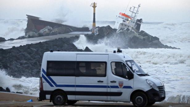 Ein gestrandetes Schiff liegt auf Felsen im Meer, während ein Polizeiwagen im Vordergrund steht.