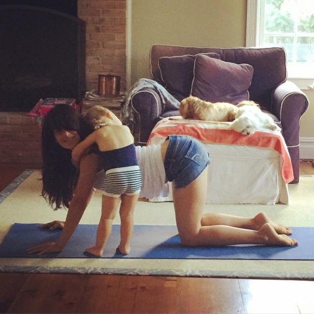 Eine Frau macht Yoga auf einer Matte, während ein kleines Kind sie umarmt. Im Hintergrund liegen zwei Hunde auf einem Sessel.